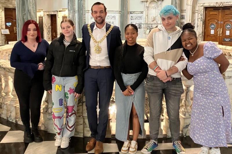 four teenage girls and one teenage boy standing beside a man with dark hair and beard, wearing suit - with marble floors and pillars behind the group